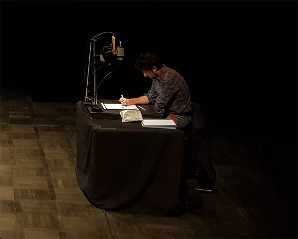 Photo d'un artiste assis sur une chaise, avec un banc titre sur la table devant lui, en train de dessiner.
